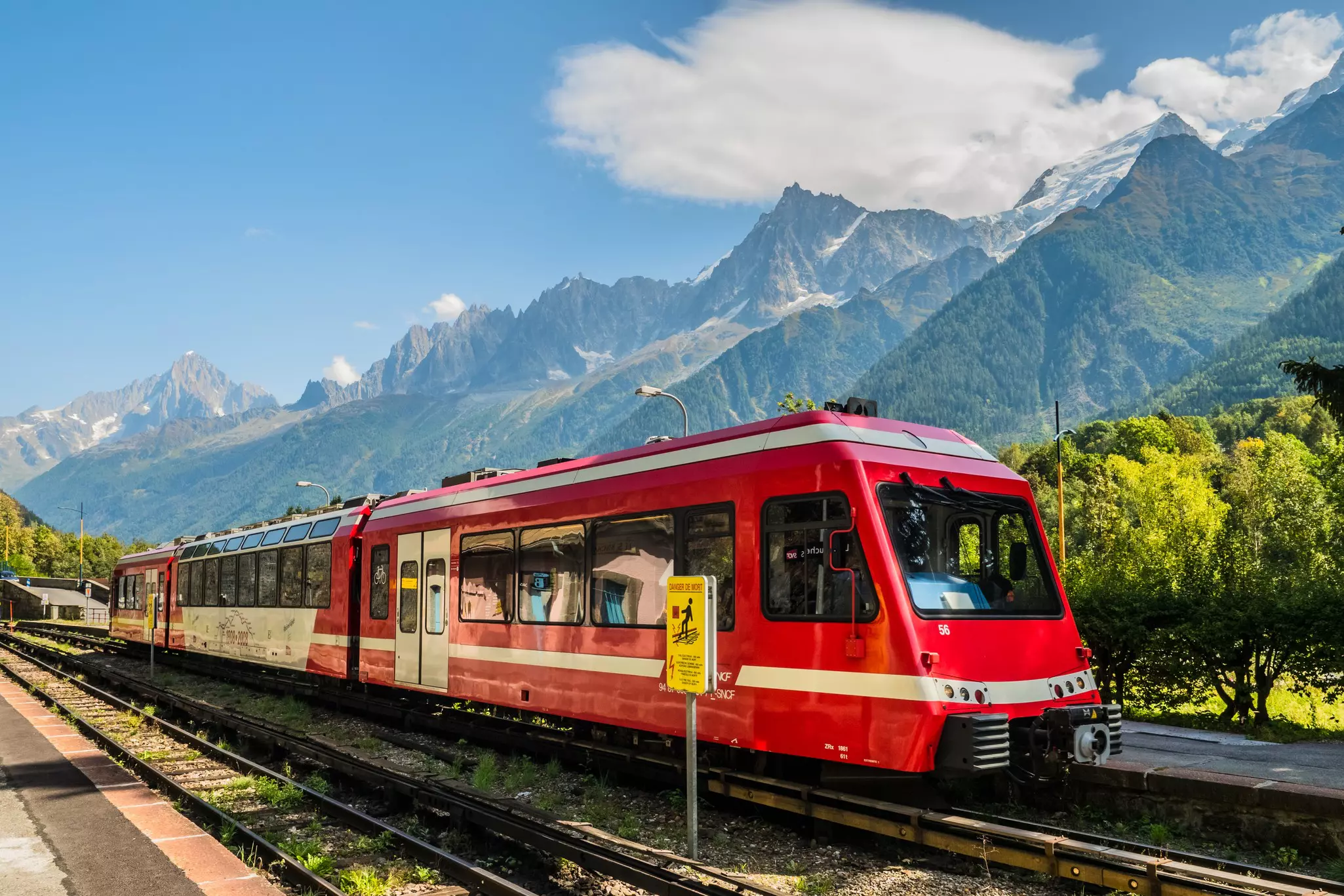 A train with three red cars travels on a track in a mountain valley with peaks rising in the distance.