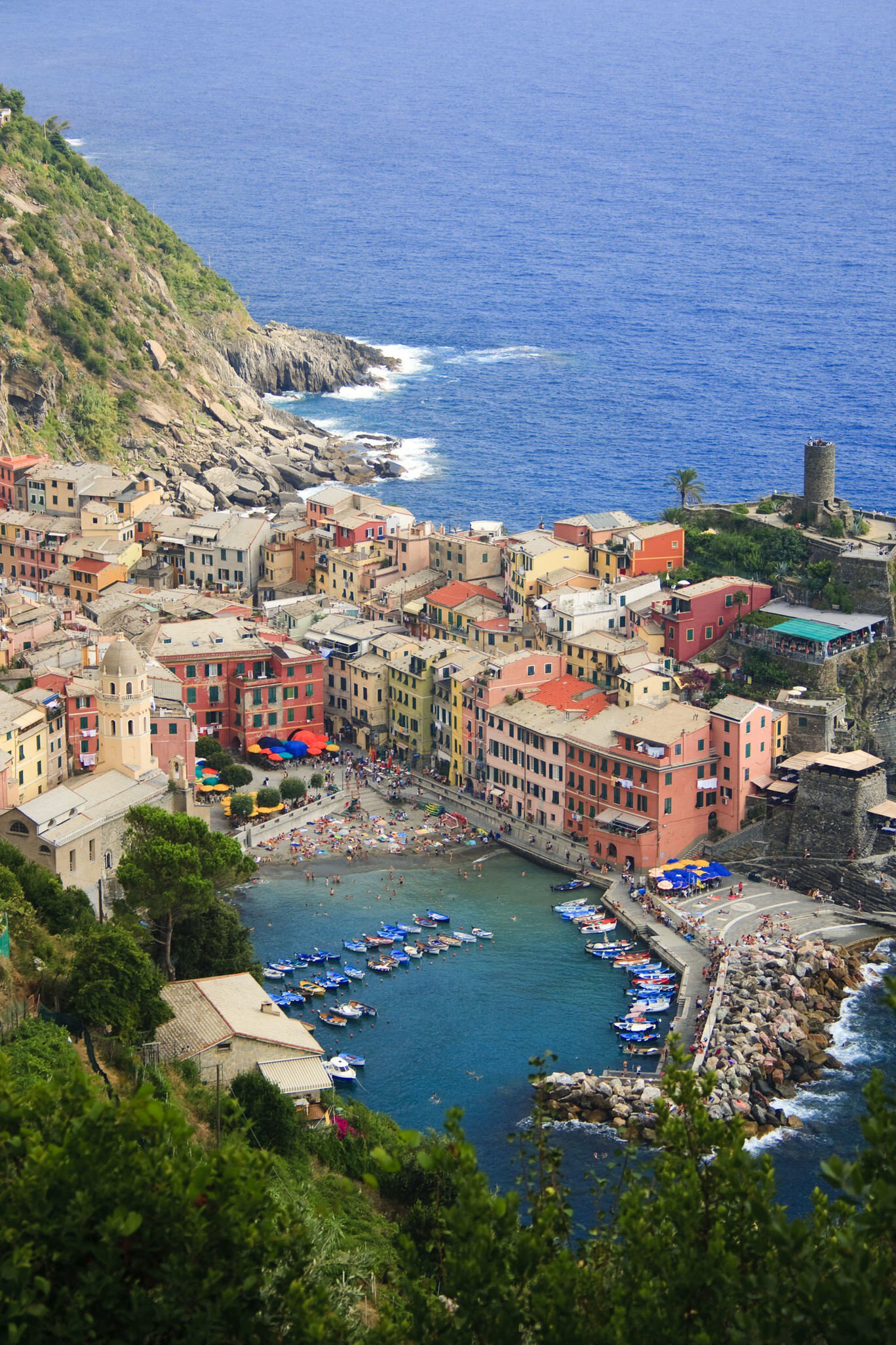 Colourful village houses around a small harbour with moored boats and a stone tower, beside a steep green headland and sea.