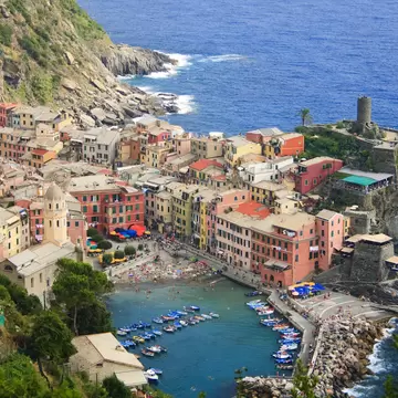 Colourful village houses around a small harbour with moored boats and a stone tower, beside a steep green headland and sea.