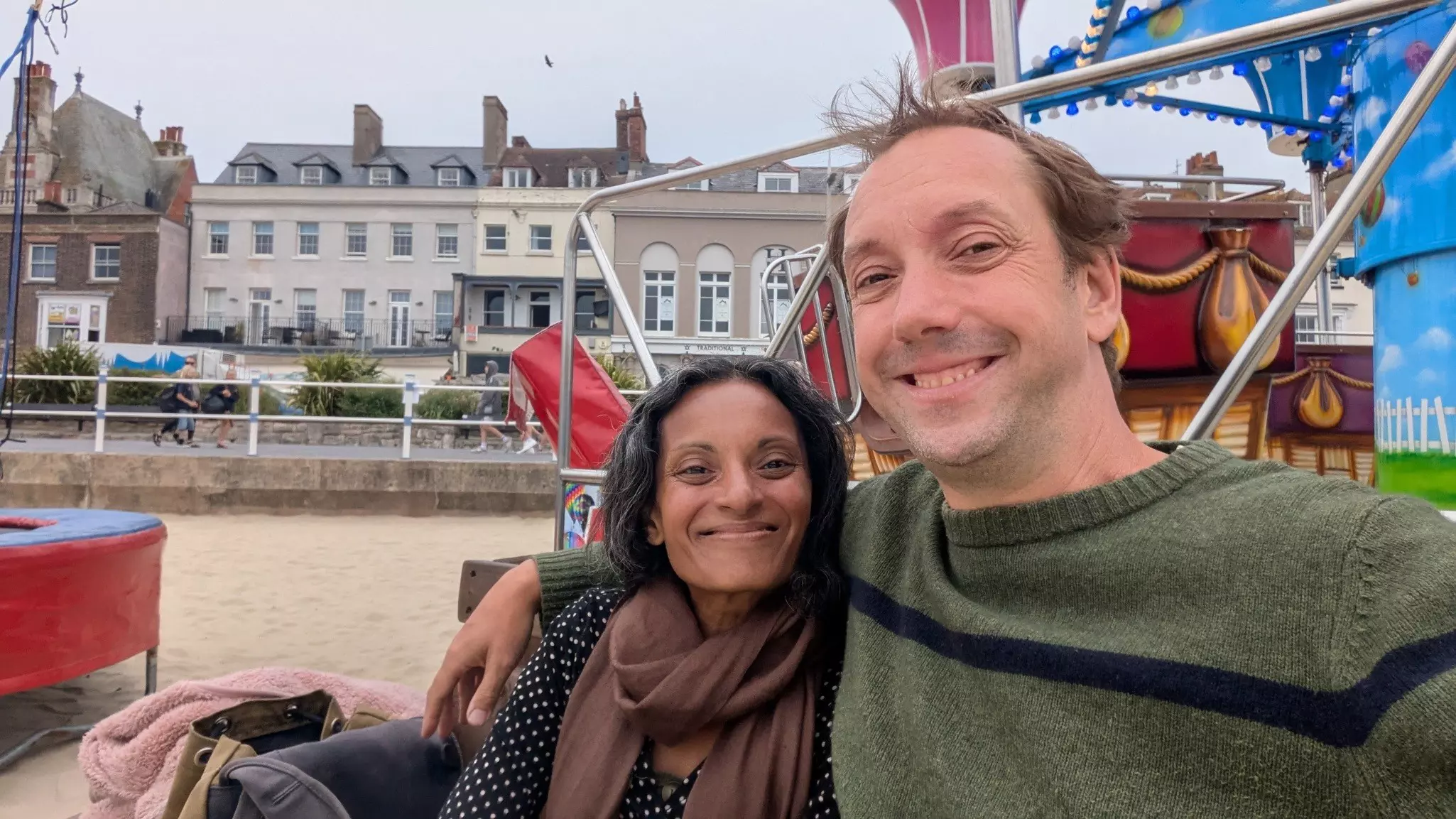 A man in a green sweater has his arm around a woman with a brown scarf on a beach in front of a variety of buildings.