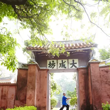 Man in a blue shirt doing tai chi in a red brick gateway with a tiled roof and Chinese characters, under branches, Tainan.