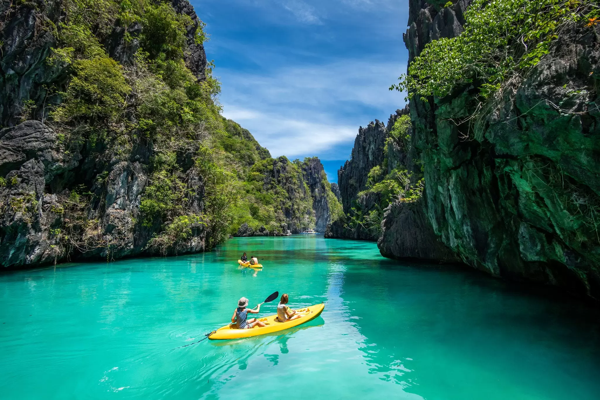Tourists on yellow kayaks going through green-blue waters bordered by craggy peaks
