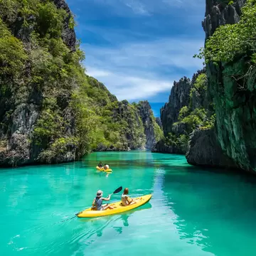 Tourists on yellow kayaks going through green-blue waters bordered by craggy peaks