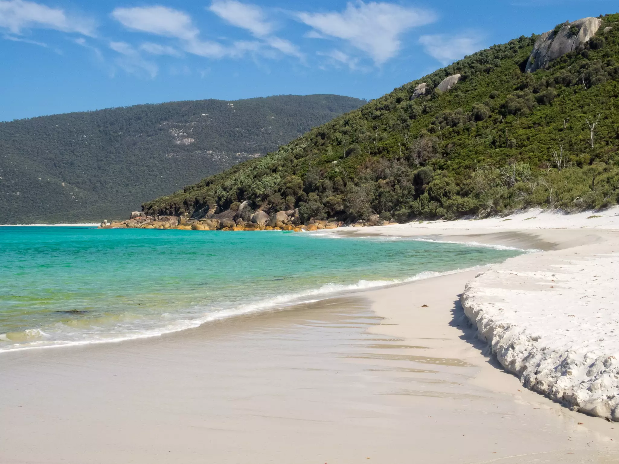 A sandy bay abuts a turquoise ocean with green tree-covered hills in the background