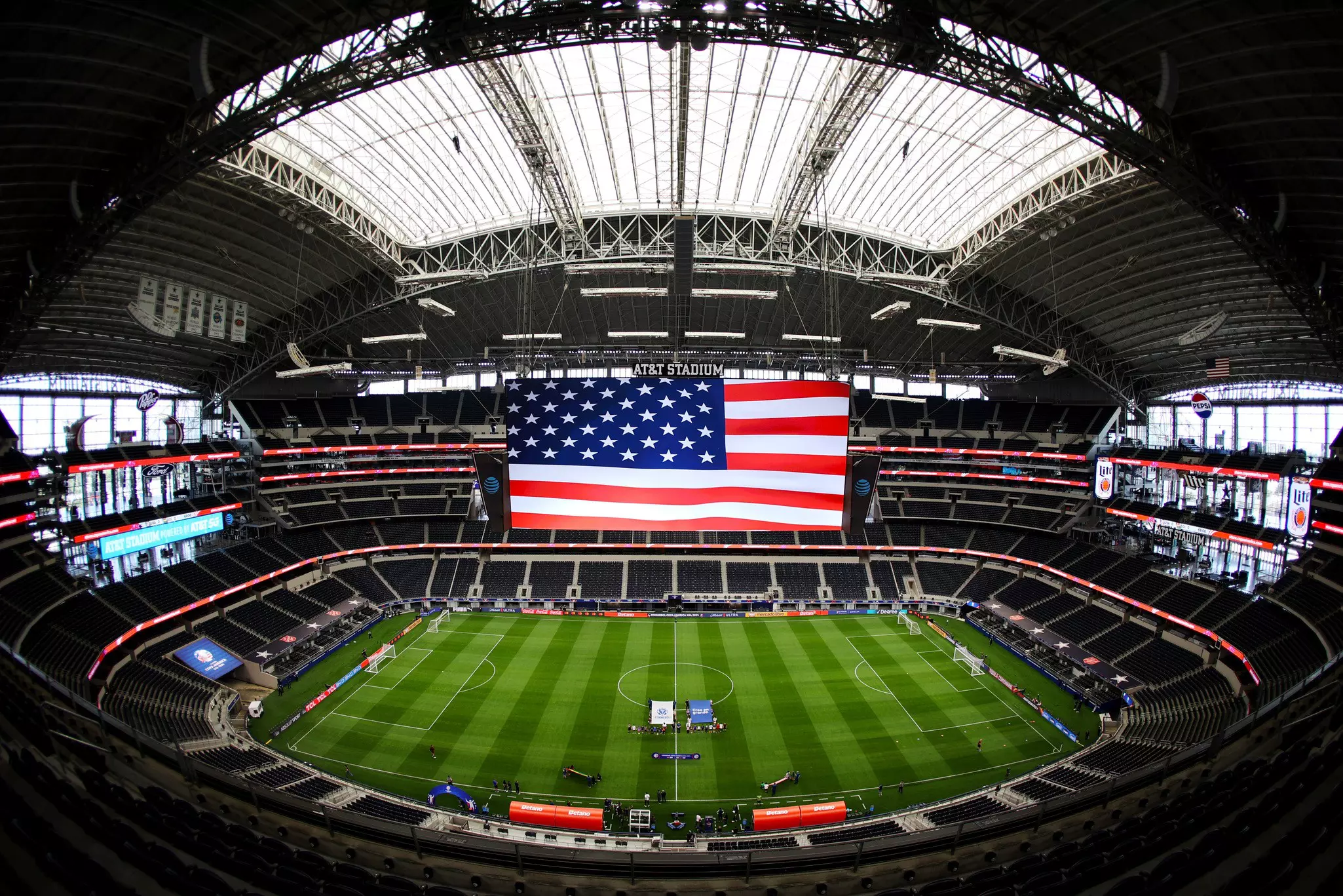 A fish-eye view of a huge stadium with a green soccer pitch. An American flag is hoisted over the field.