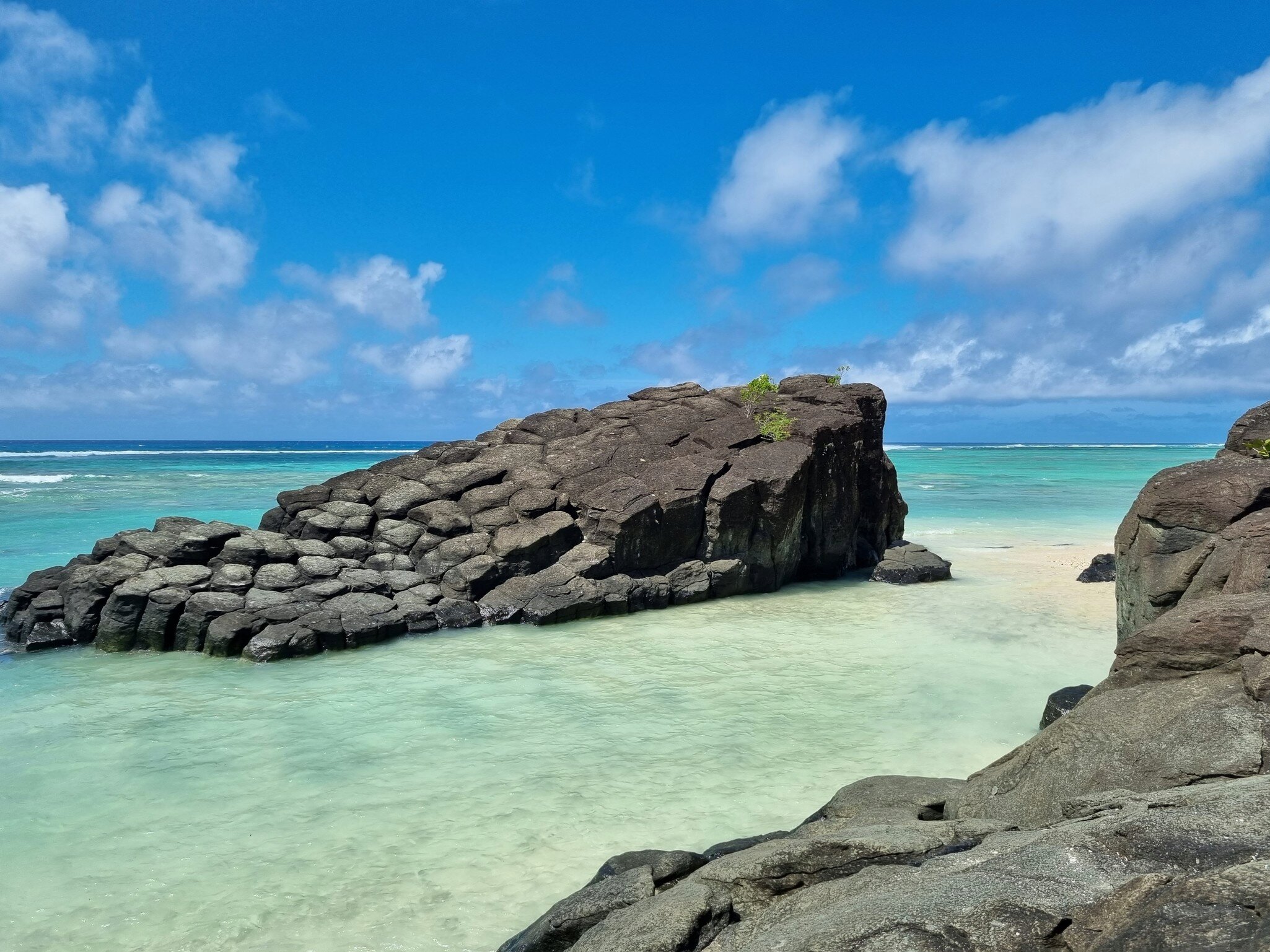 Dark columnar rock rising from shallow turquoise water under a blue sky with white clouds