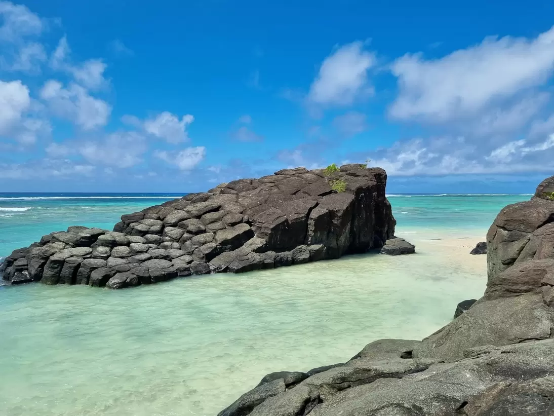Dark columnar rock rising from shallow turquoise water under a blue sky with white clouds