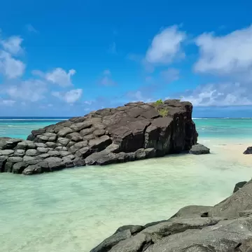 Dark columnar rock rising from shallow turquoise water under a blue sky with white clouds