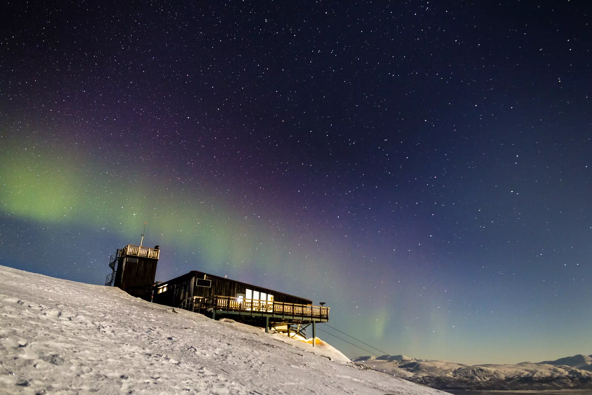 The northern lights and stars shine above a wooden building perched atop a snowy hill.