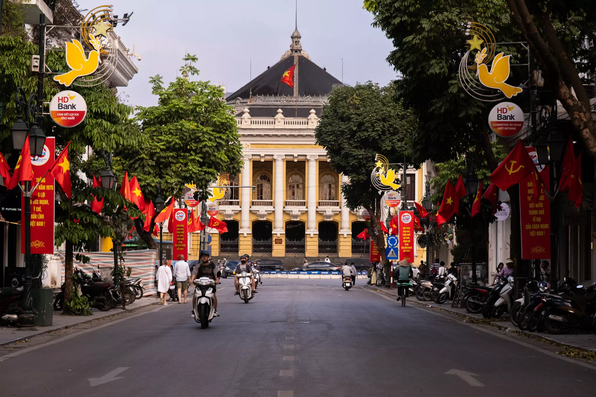 Motorcyclists ride by a large columned building in Hanoi, Vietnam.