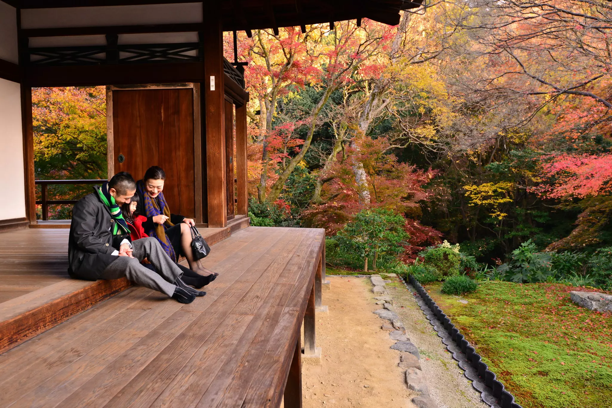 Two parents sit with their child on the terrace of a wooden temple. Fall foliage can be seen in the woods beyond.