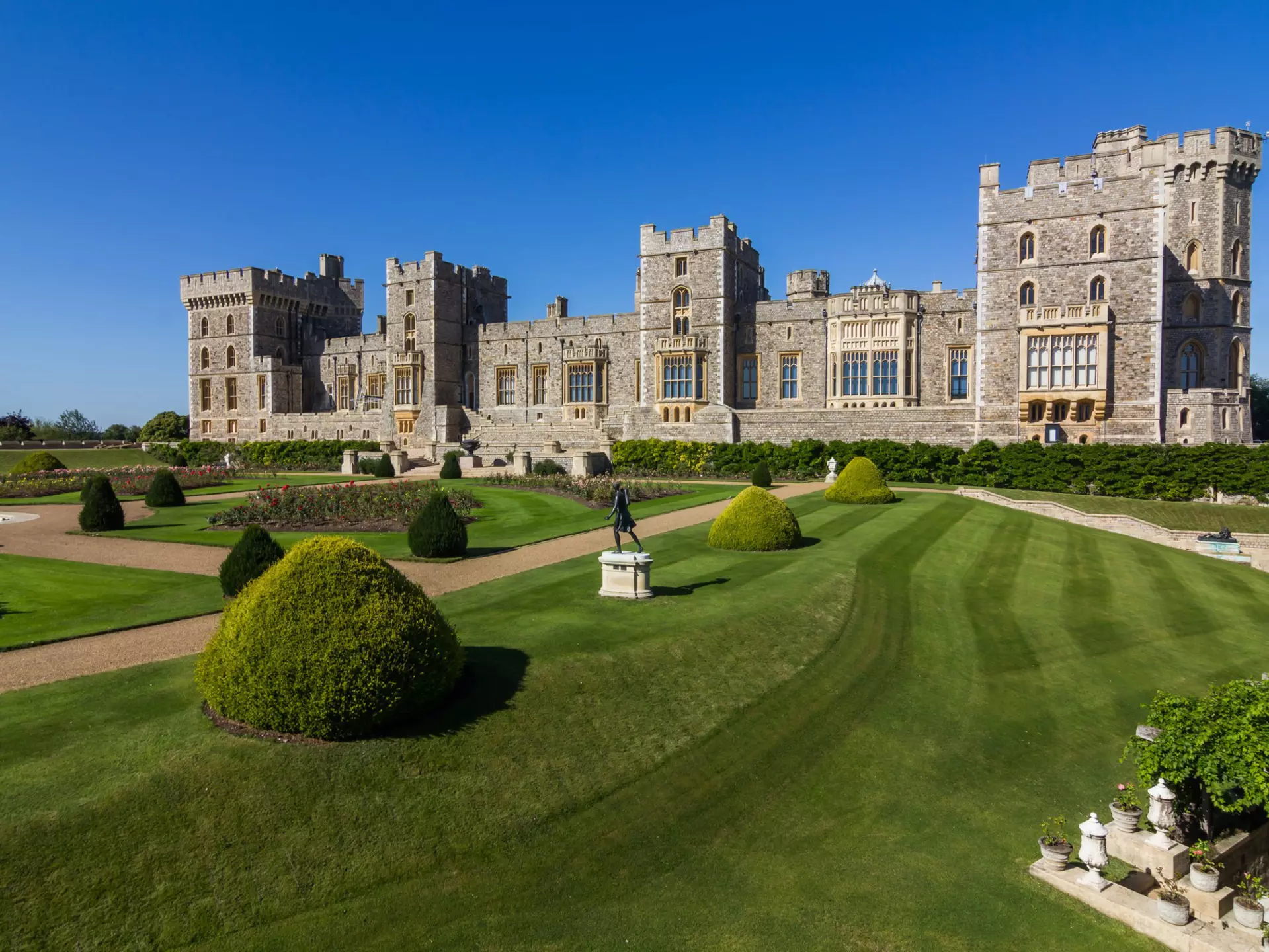 Windsor Castle's grey stone walls and castellated towers rise into a clear blue sky, with the extensive manicured lawn, hedges and shrubs layed out in the foreground © Kanuman / Shutterstock