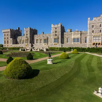 Windsor Castle's grey stone walls and castellated towers rise into a clear blue sky, with the extensive manicured lawn, hedges and shrubs layed out in the foreground © Kanuman / Shutterstock