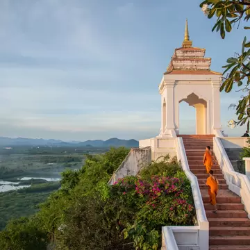 Two monks in saffron robes walking up stairs to a hilltop shrine, with bougainvillea and a green plain below