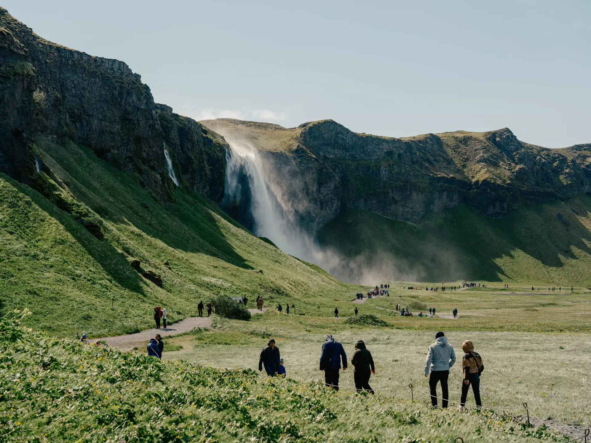 Individual groups of hikers walk along a green path towards a waterfall on a sunny day