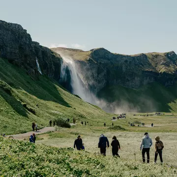 Individual groups of hikers walk along a green path towards a waterfall on a sunny day