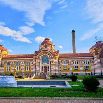 A large striped-brick building with domed pavilions and glazed ceramic ornament, fronted by a shallow pool and lawns.