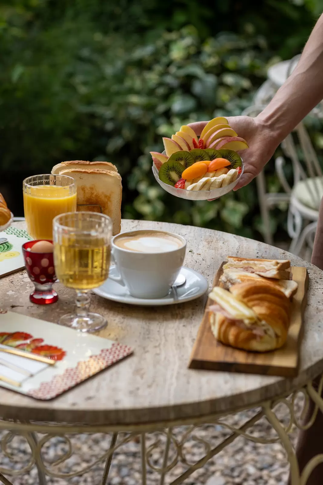 A table in a garden with croissants, a hard-boiled egg, a latte, orange juice, toast, and a hand holding a bowl of fruit