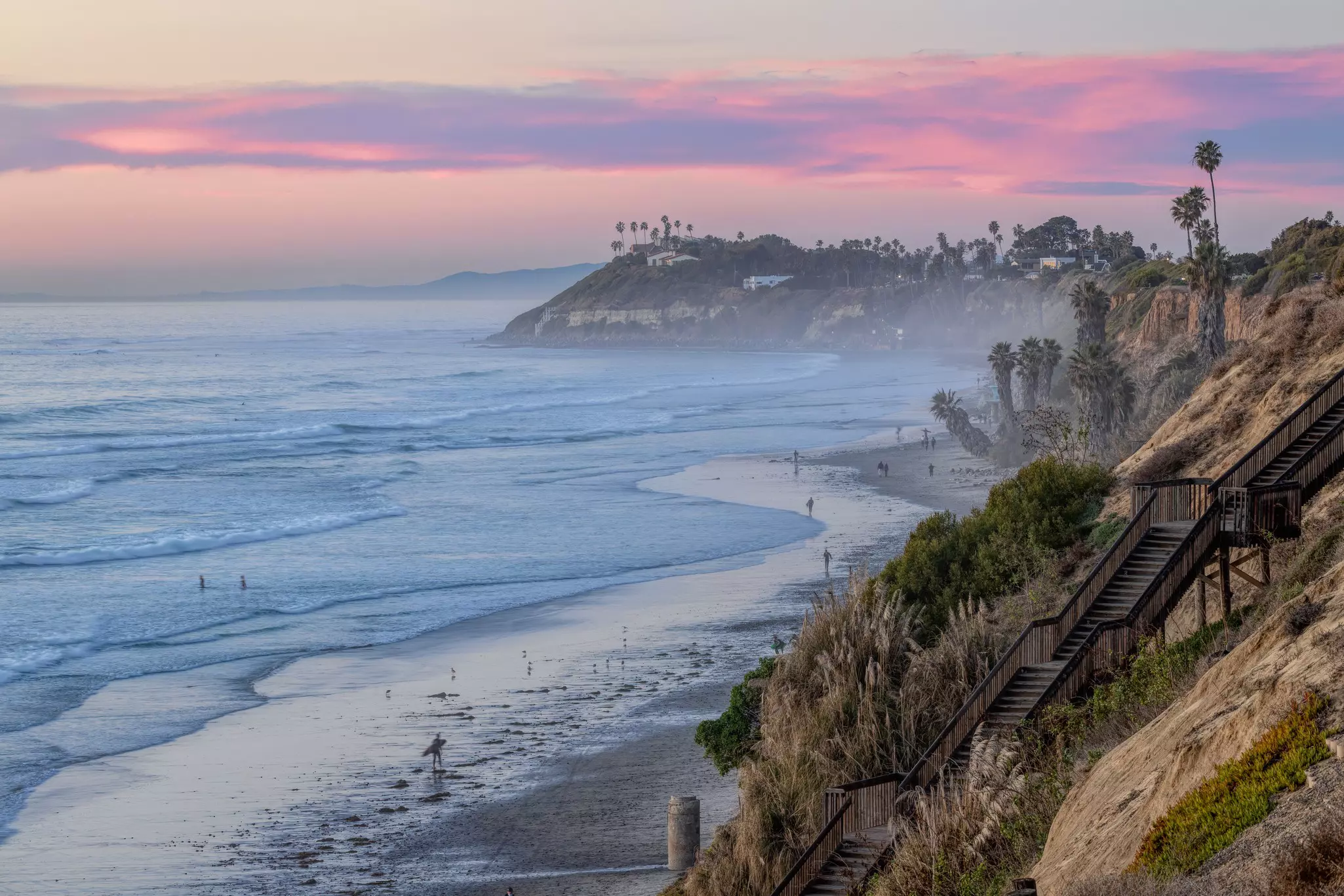 A beach at dawn with surfers out in the waves as a pink light is cast over the sky.