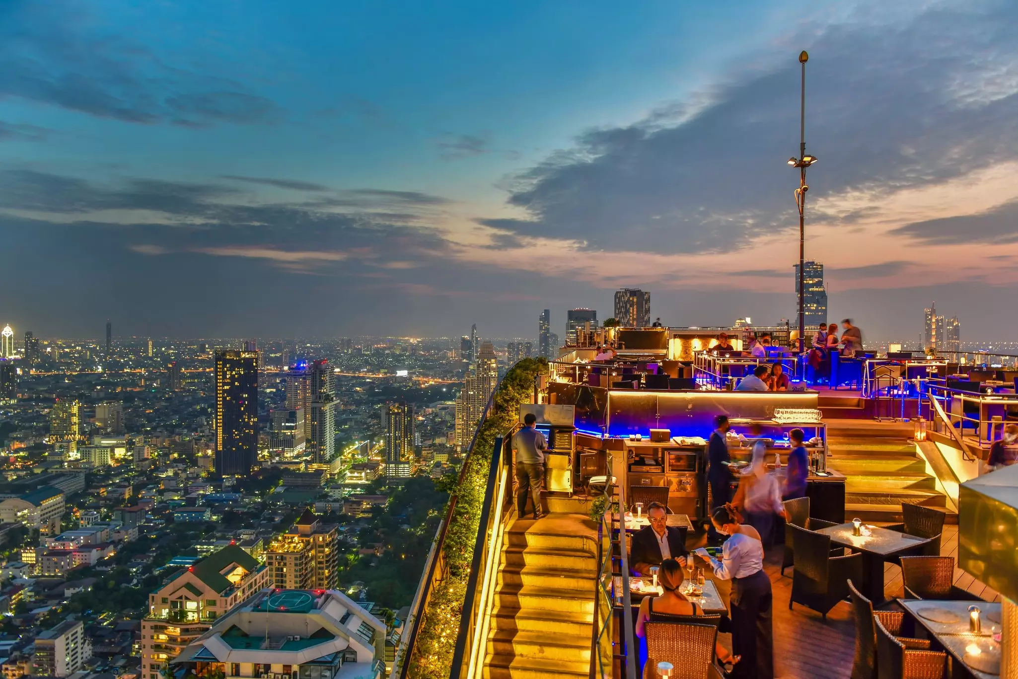 A view of a rooftop bar overlooking the skyline of a huge city at dusk.