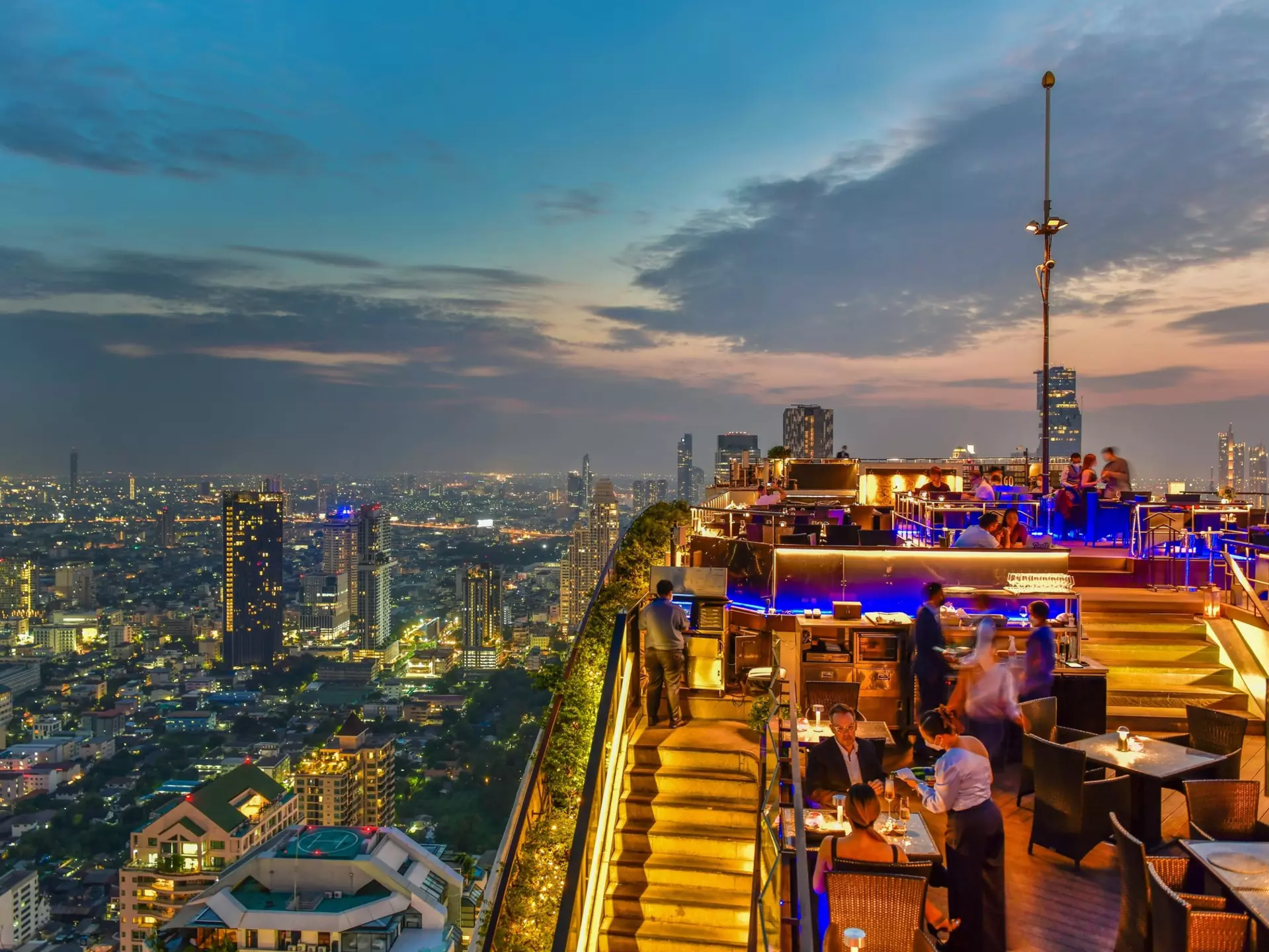 A view of a rooftop bar overlooking the skyline of a huge city at dusk.