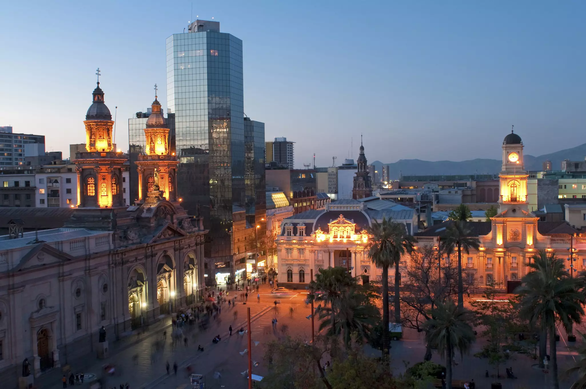 An overhead view of a city square at dusk, with historic buildings lining the palm-filled plaza.
