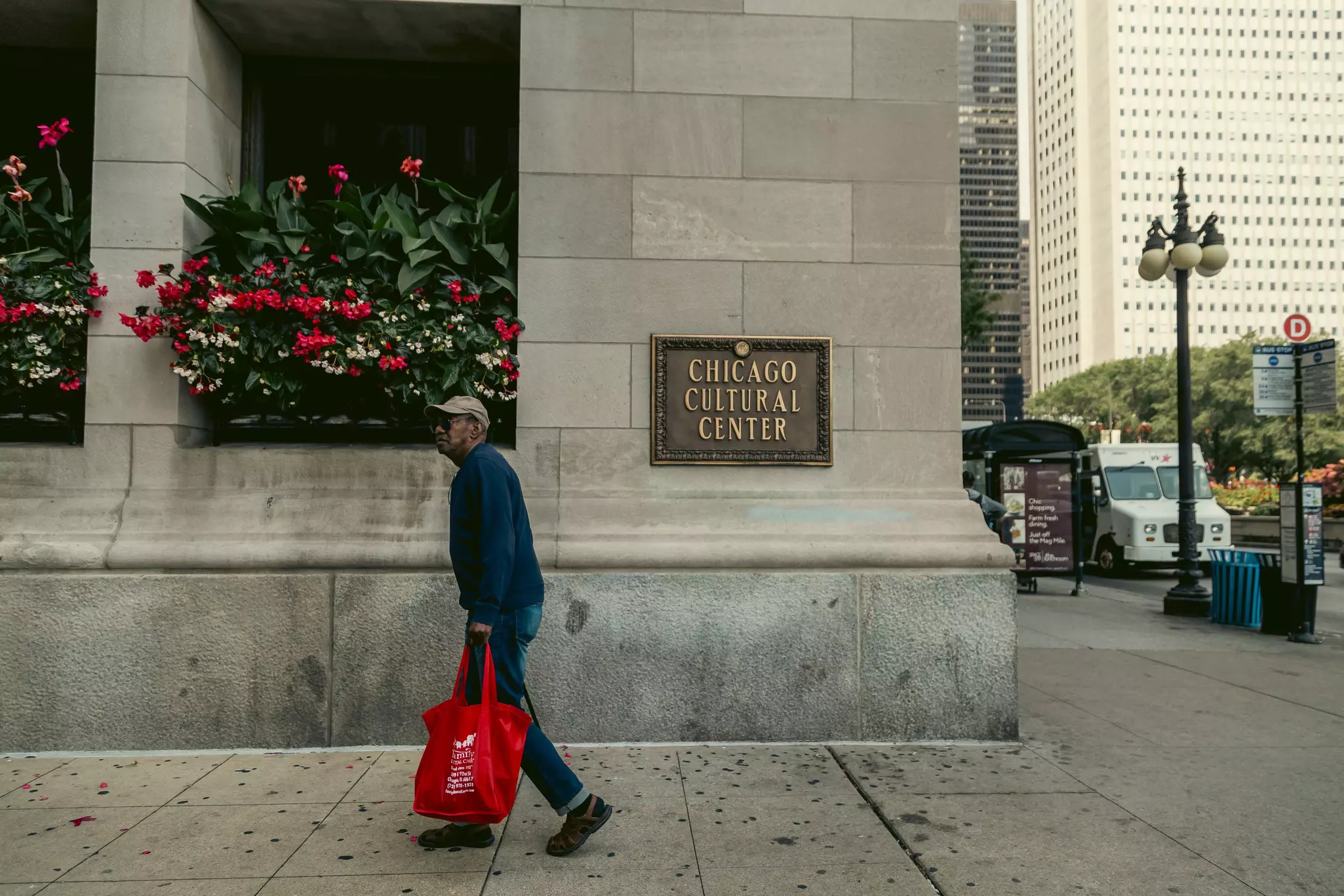 A man holding a red tote bag walks on the sidewalk of a city past a large institutional building.