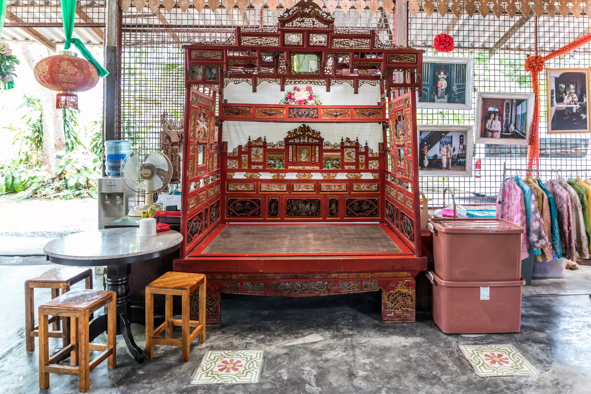 A red, altarlike structure with ornate decorations. A table fan is on the left side of the photo, and two storage bins are on the right