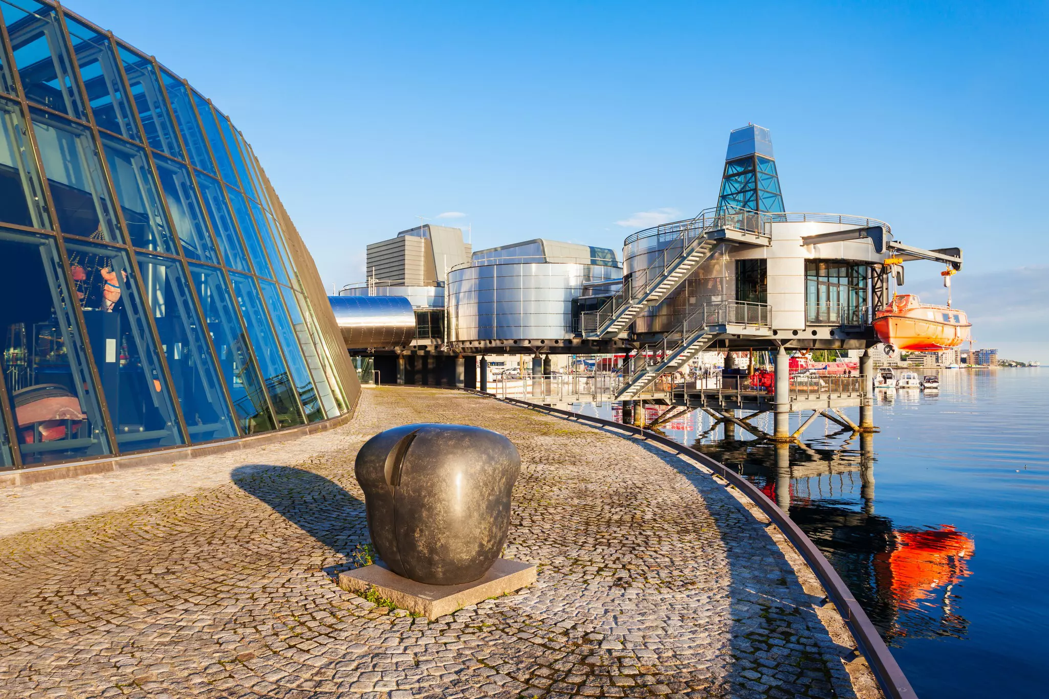 A cobbled waterfront pathway leads to a museum constructed of metal-clad buildings built over the water, in Stavanger, Norway.