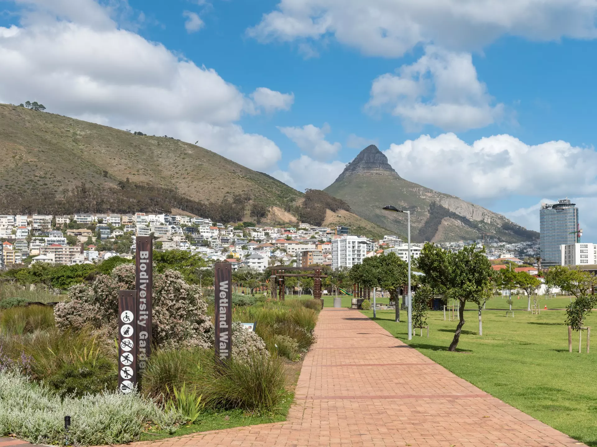 A path leads through a tidy city park. A dense neighborhood nestled against the slope of a mountain is visible in the distance.