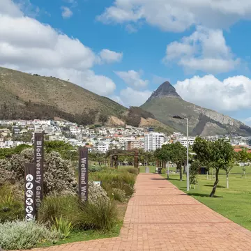 A path leads through a tidy city park. A dense neighborhood nestled against the slope of a mountain is visible in the distance.