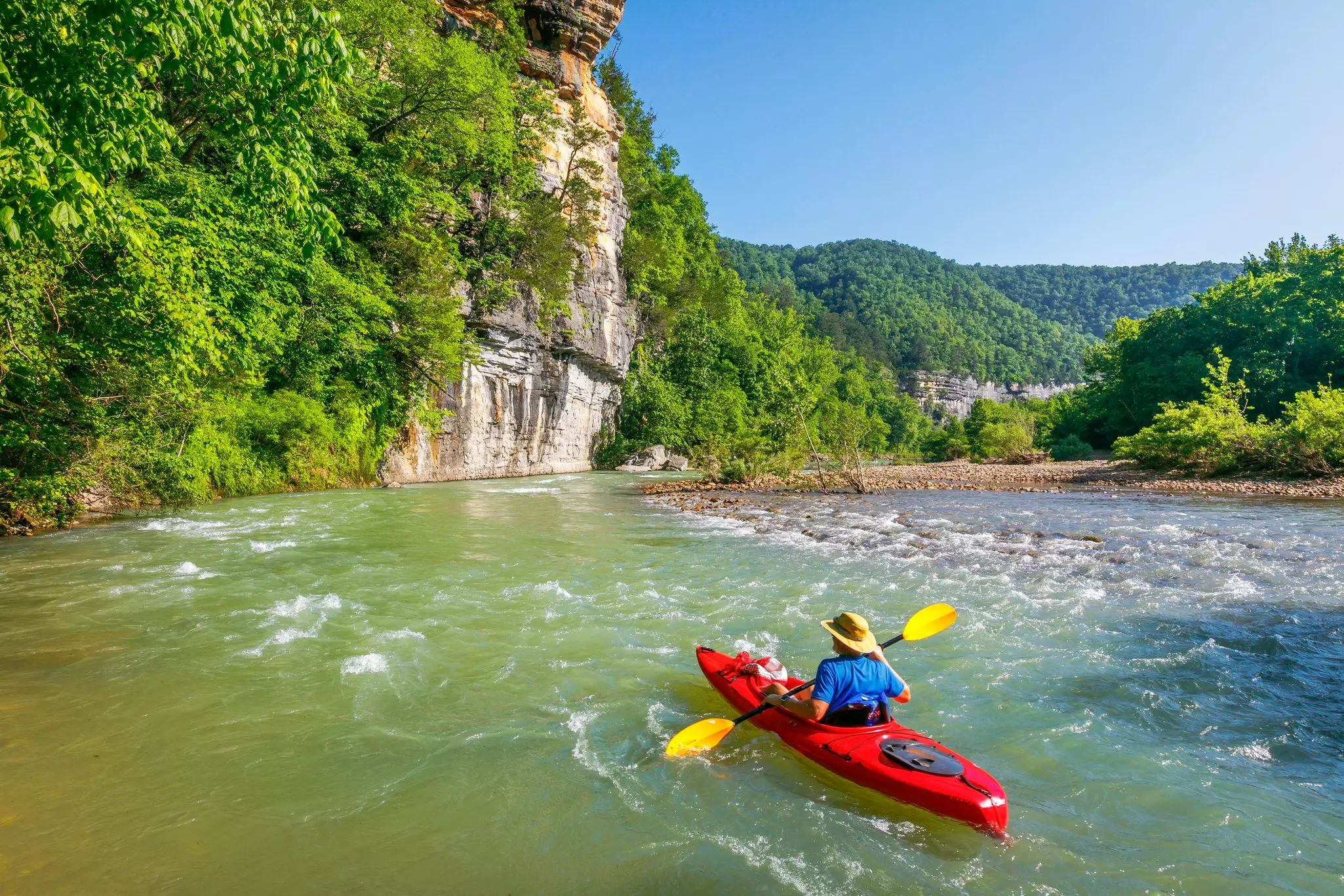 A kayaker paddles down a river past rocky cliffs.