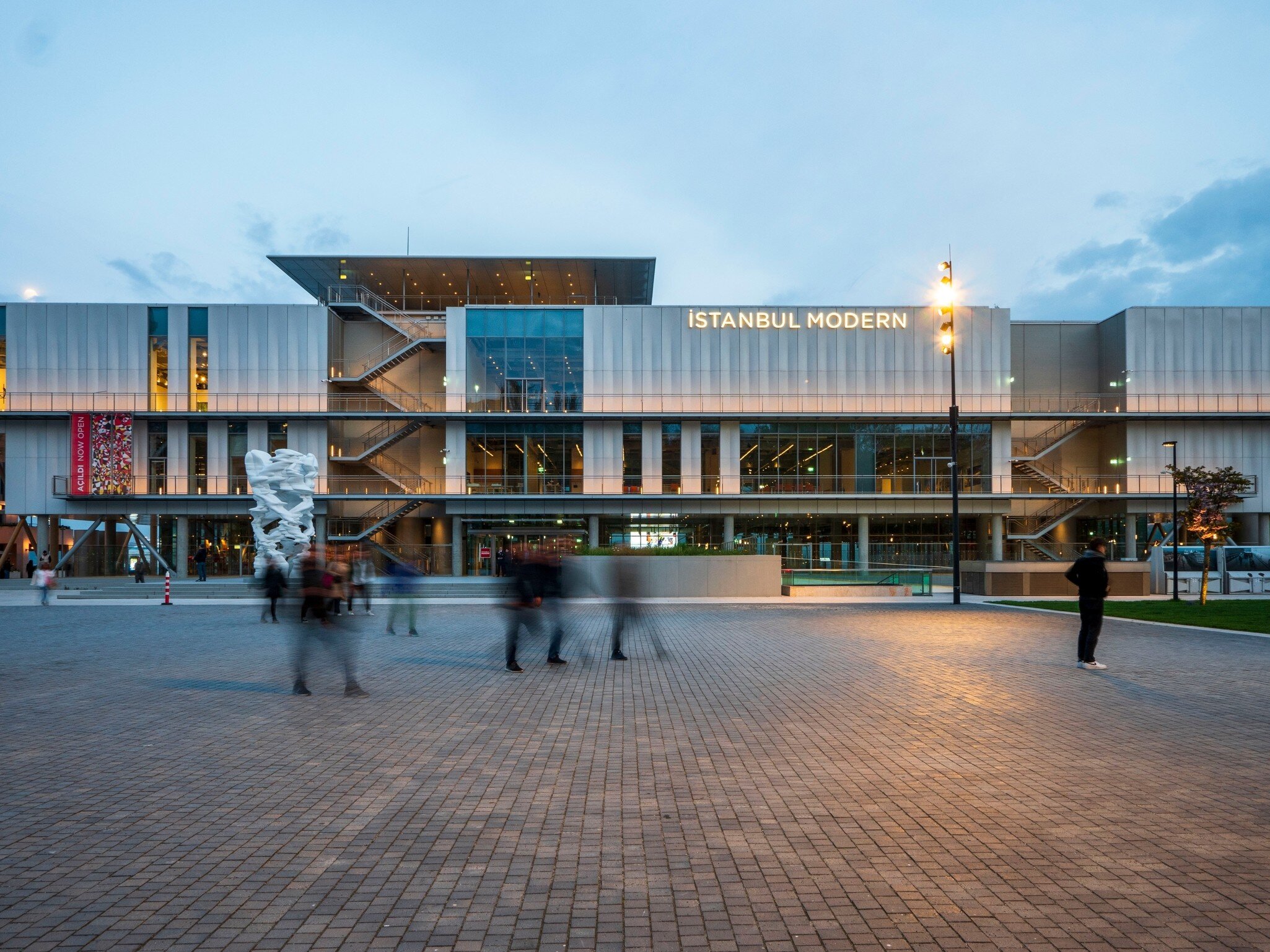 Pale panelled building with a lit name sign and external stairways above a brick plaza, people walking past at dusk