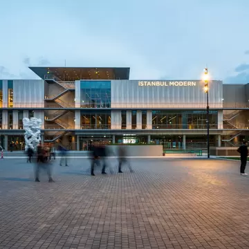 Pale panelled building with a lit name sign and external stairways above a brick plaza, people walking past at dusk