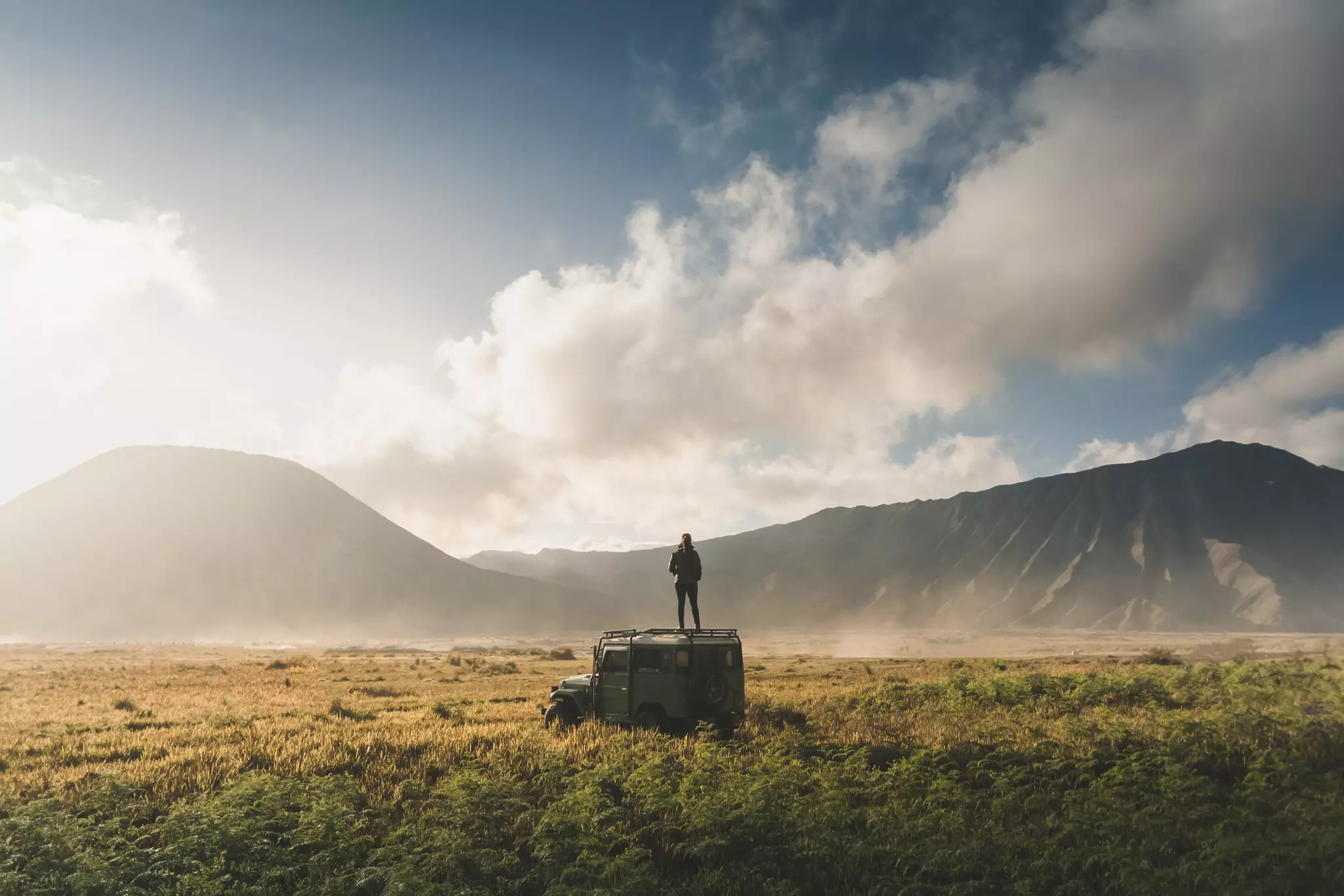 A woman stands on top of a four-wheel drive in a lava fields at the foot of a volcano.
