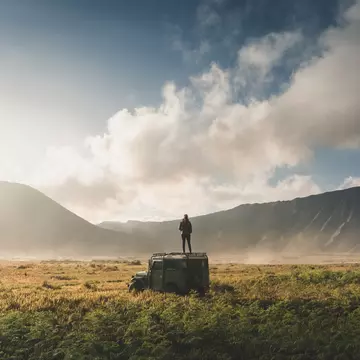 A woman stands on top of a four-wheel drive in a lava fields at the foot of a volcano.