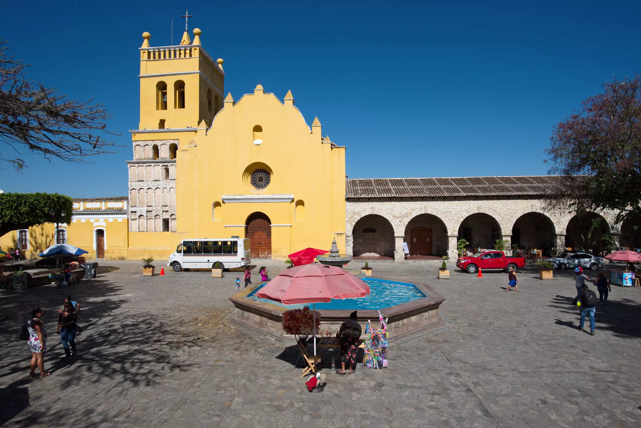 People walk and gather in a cobblestone square flanked by a large yellow church on a sunny day.