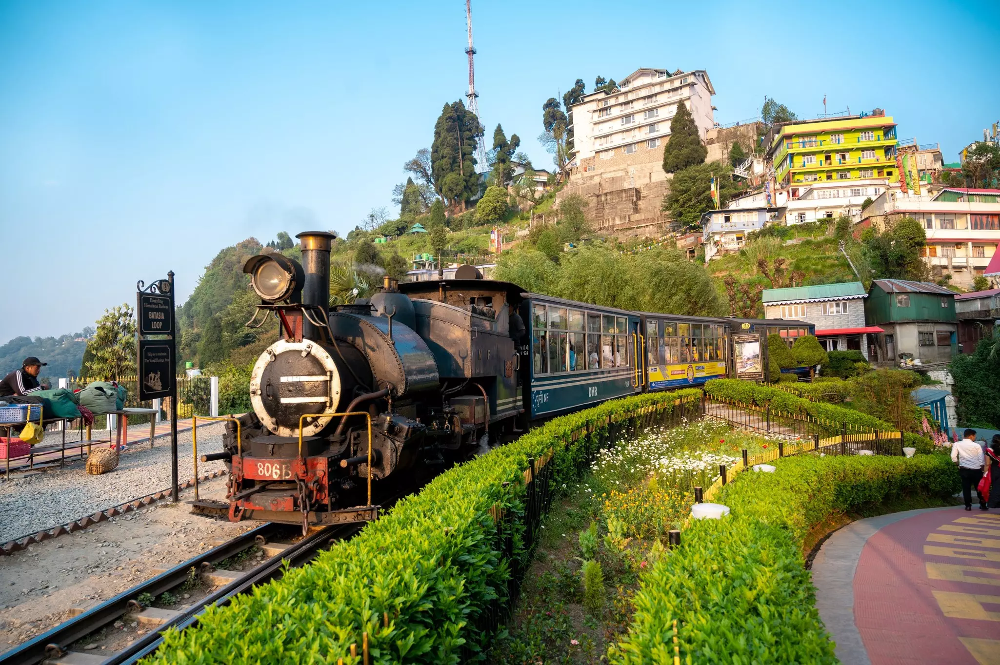 A small-gauge train is pictured on a track at the top of a hill. Buildings with balconies are seen behind the train.