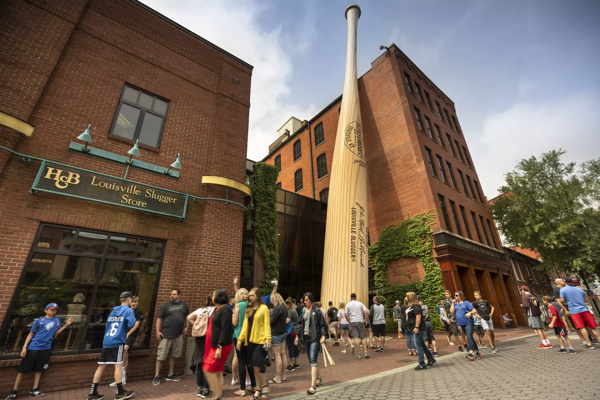 A group of people father outside a brick building. A giant baseball bat leans against the five-story building.