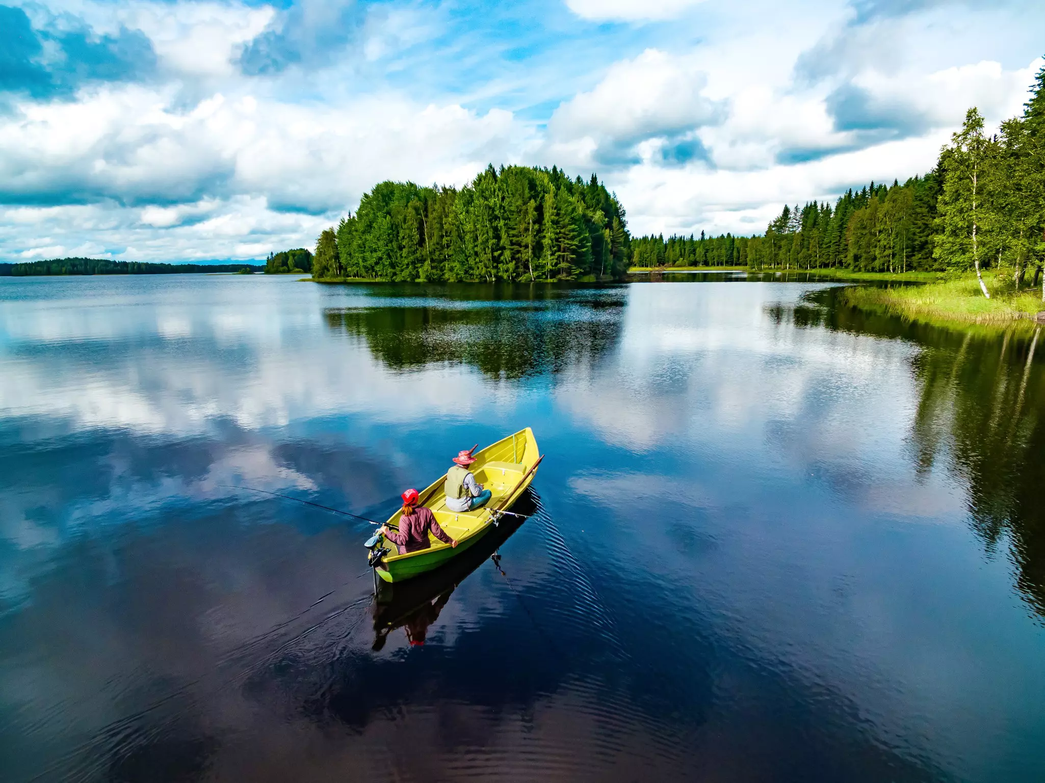 Aerial view of a yellow-painted fishing boat with young woman and man in blue lake in summer. Pine trees are seen along the coast, while the sky is partly clouded and reflected on the water