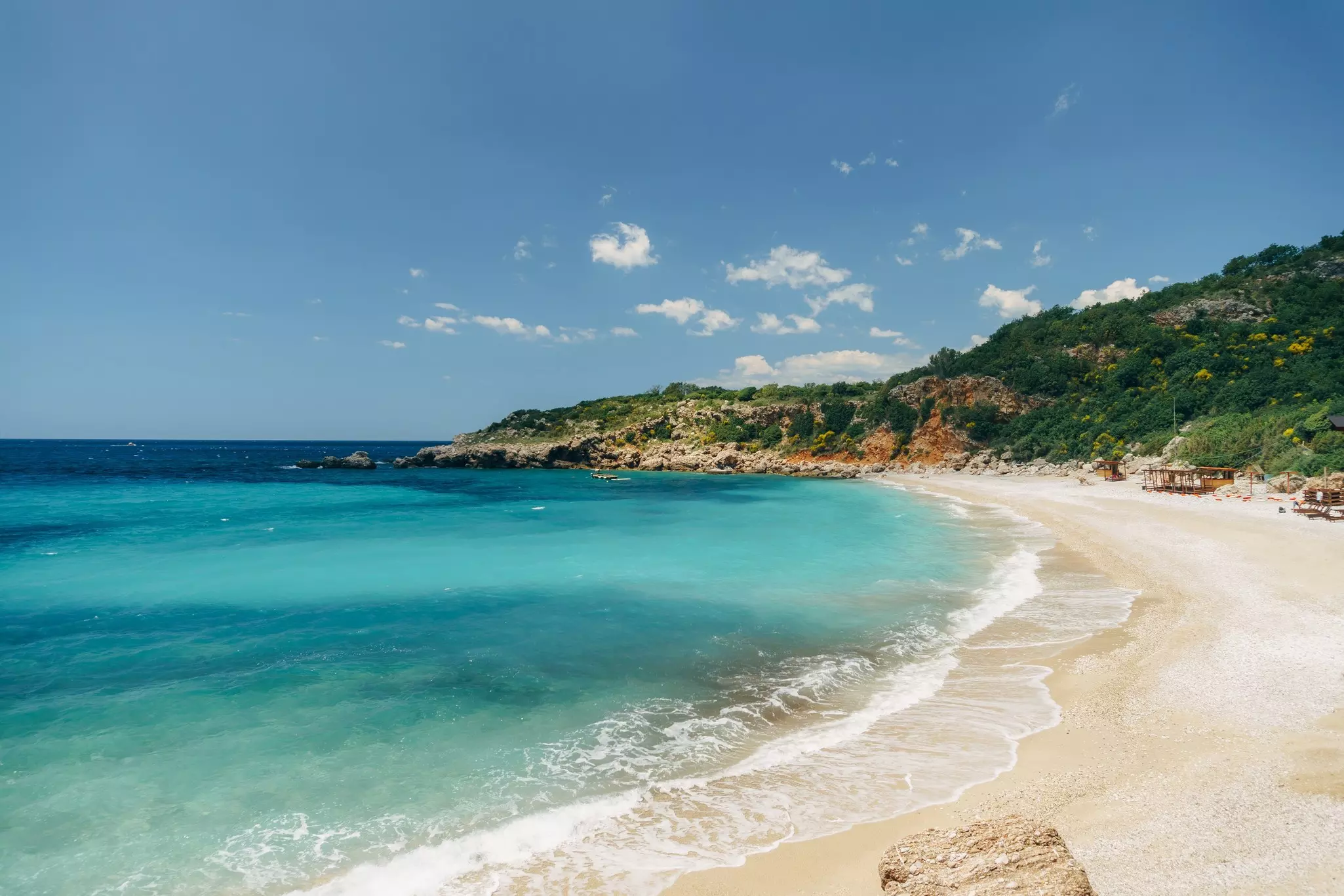 Waves lap the sand at a deserted beach. Greenery covers a hillside leading down to the beach.