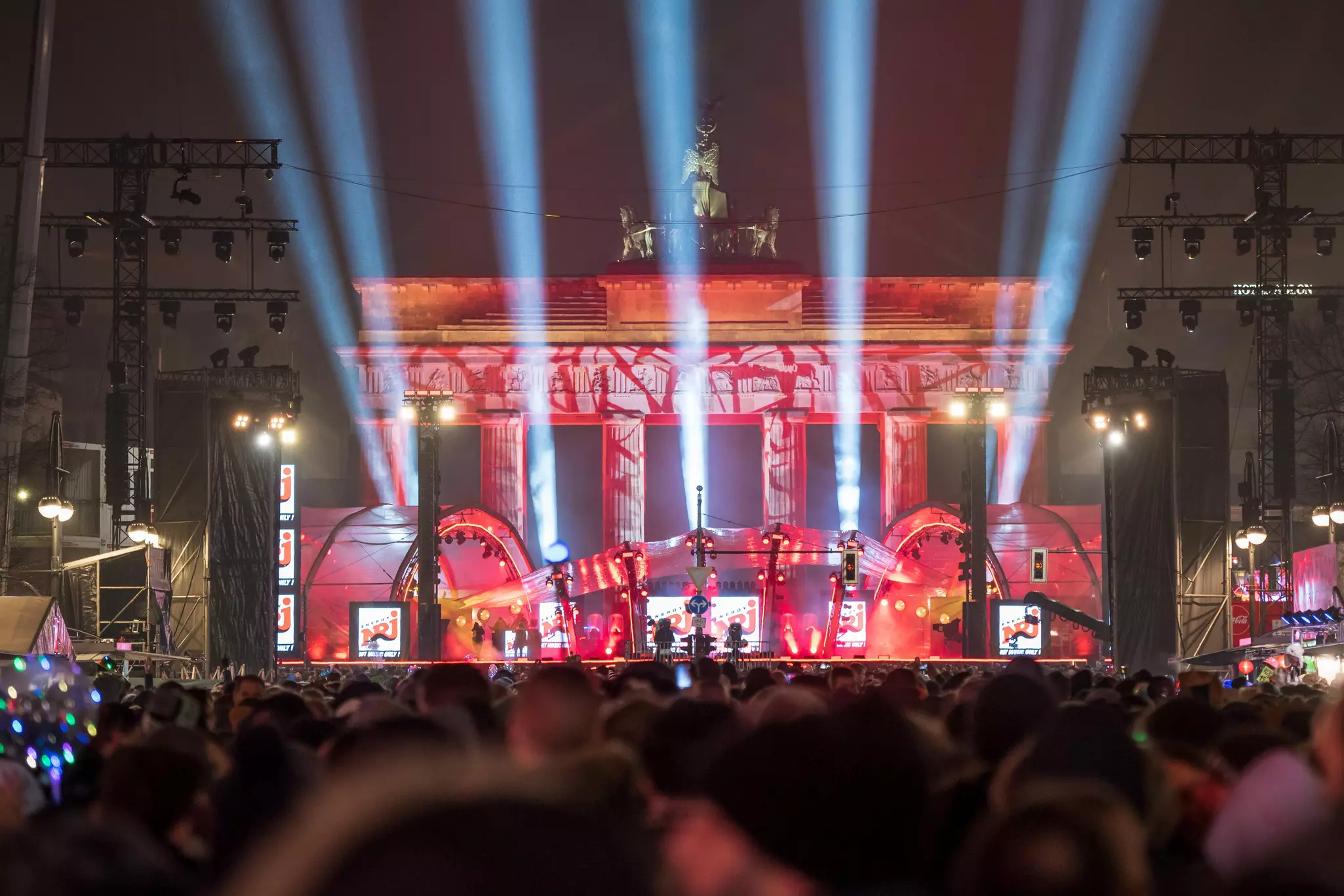 A crowd in front of a concert stage. Lights are shooting from stage and a stone monument with pillars is behind it.