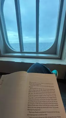 A notebook on a person's lap as they sit in a chair inside a cruise ship overlooking the ocean through a small window.