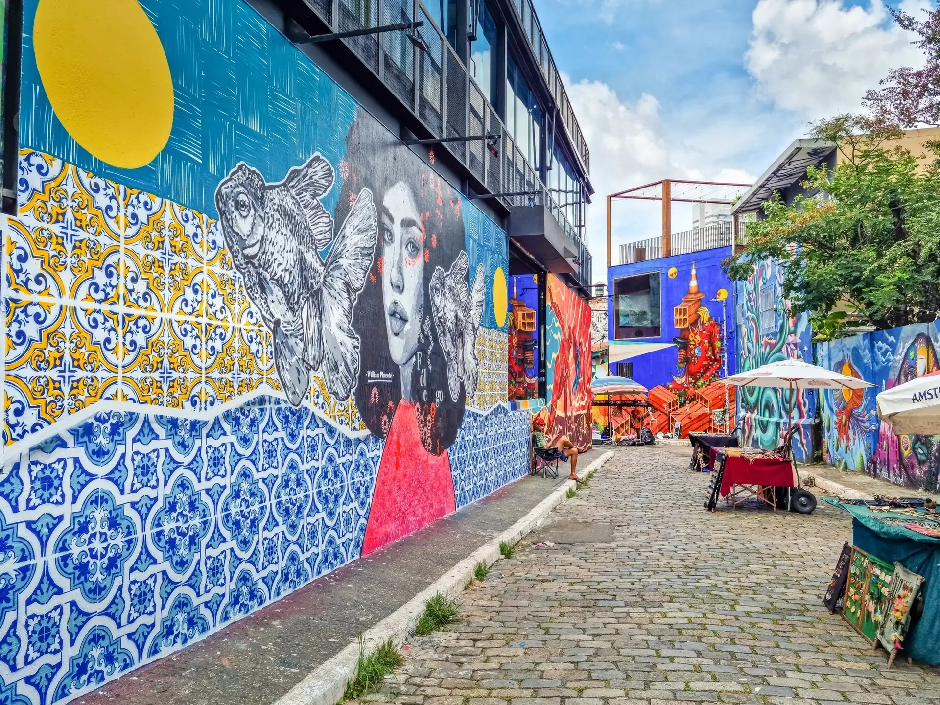 Man sitting in an alley in front of a mural of a woman's head with two fish floating by her