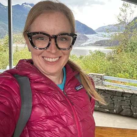 A woman wearing a red winter jacket and glasses smiles, with water and forest in the backbround.