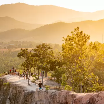 People standing on narrow sandstone ridges among pine trees at Pai Canyon, backlit by golden sunset haze over hills.