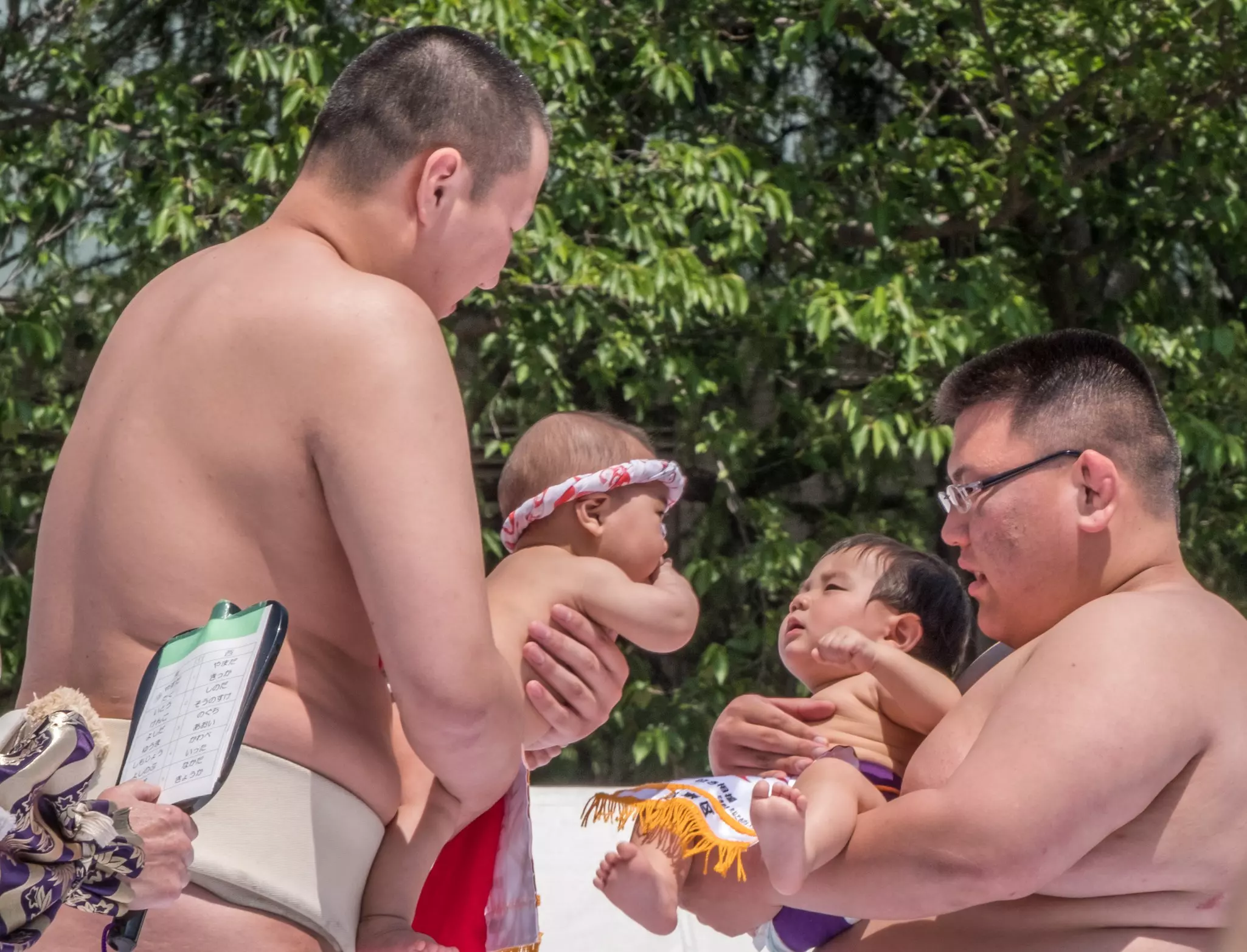Two sumo wrestlers hold babies facing each other on a sunny day.