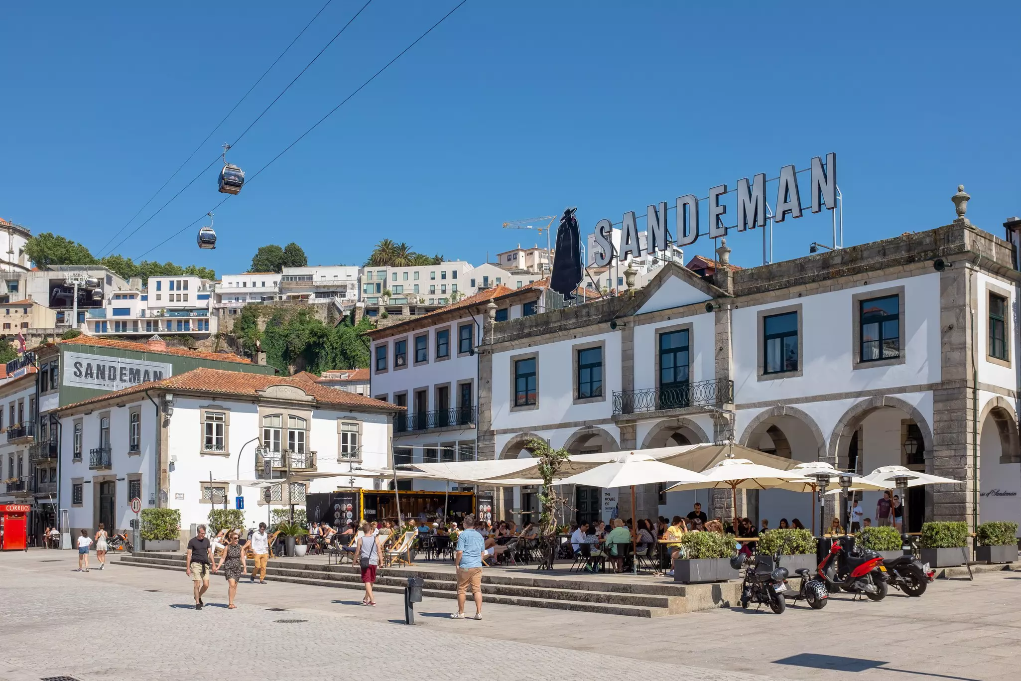 People sit at tables under white umbrellas at an outdoor patio cafe; there is a large sign for Sandeman above the building.