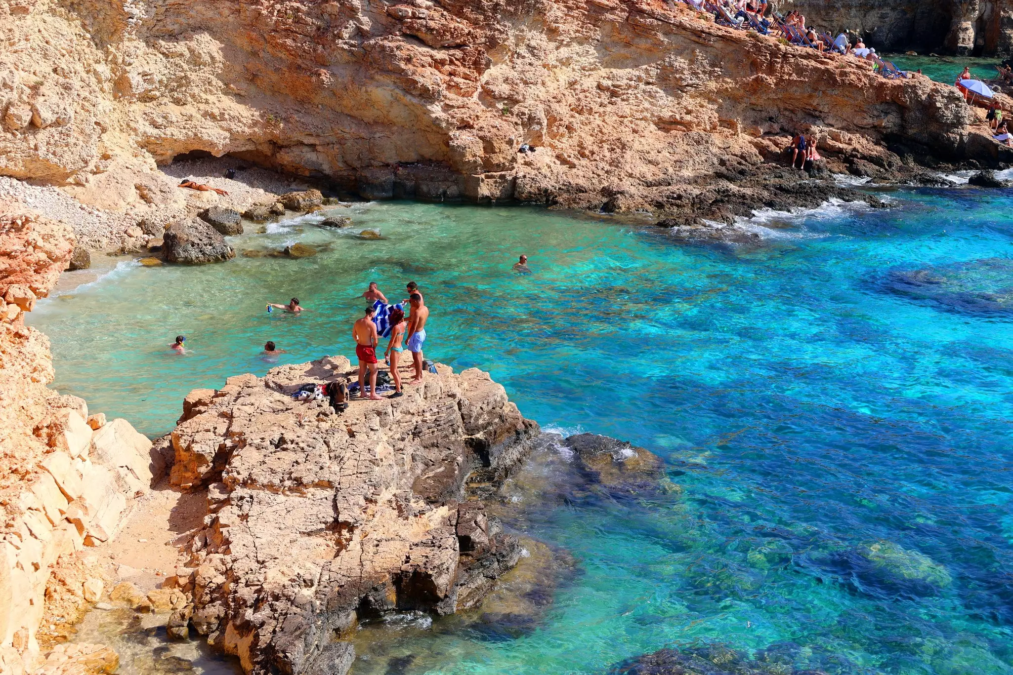 People in bathing suits stand on a rocky outcrop above a beautiful blue sheltered ocean cove
