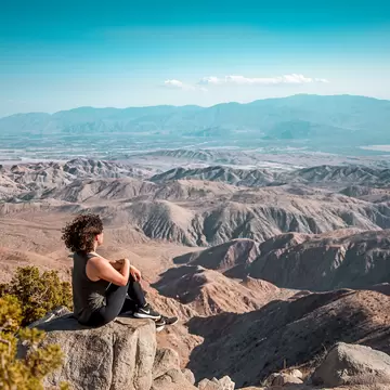 Woman with curly hair seated on a granite rock above folded brown desert hills, a hazy valley and distant mountain range